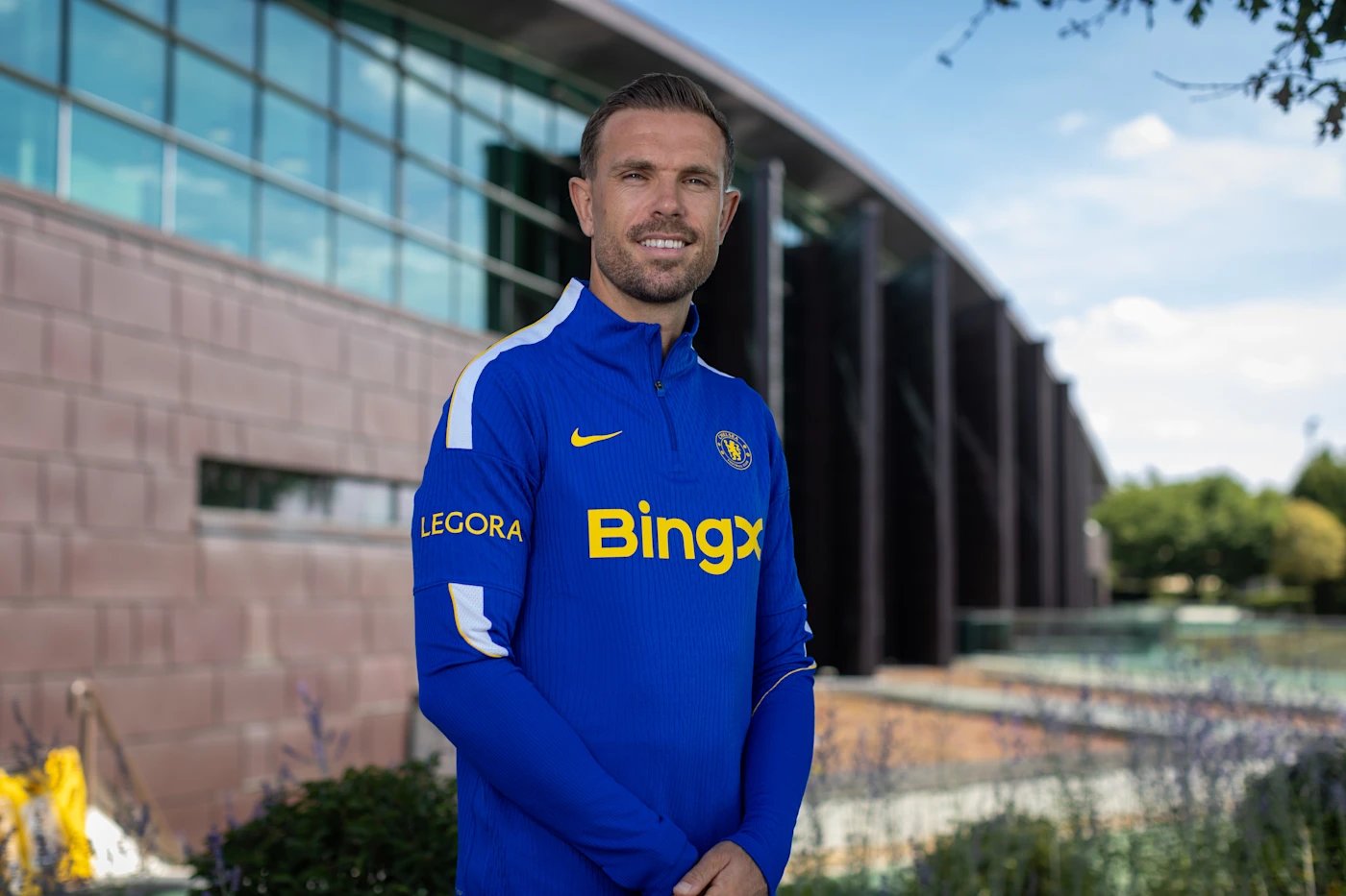 Jordan Henderson smiling in a Chelsea training top outside the club's Cobham training ground after signing